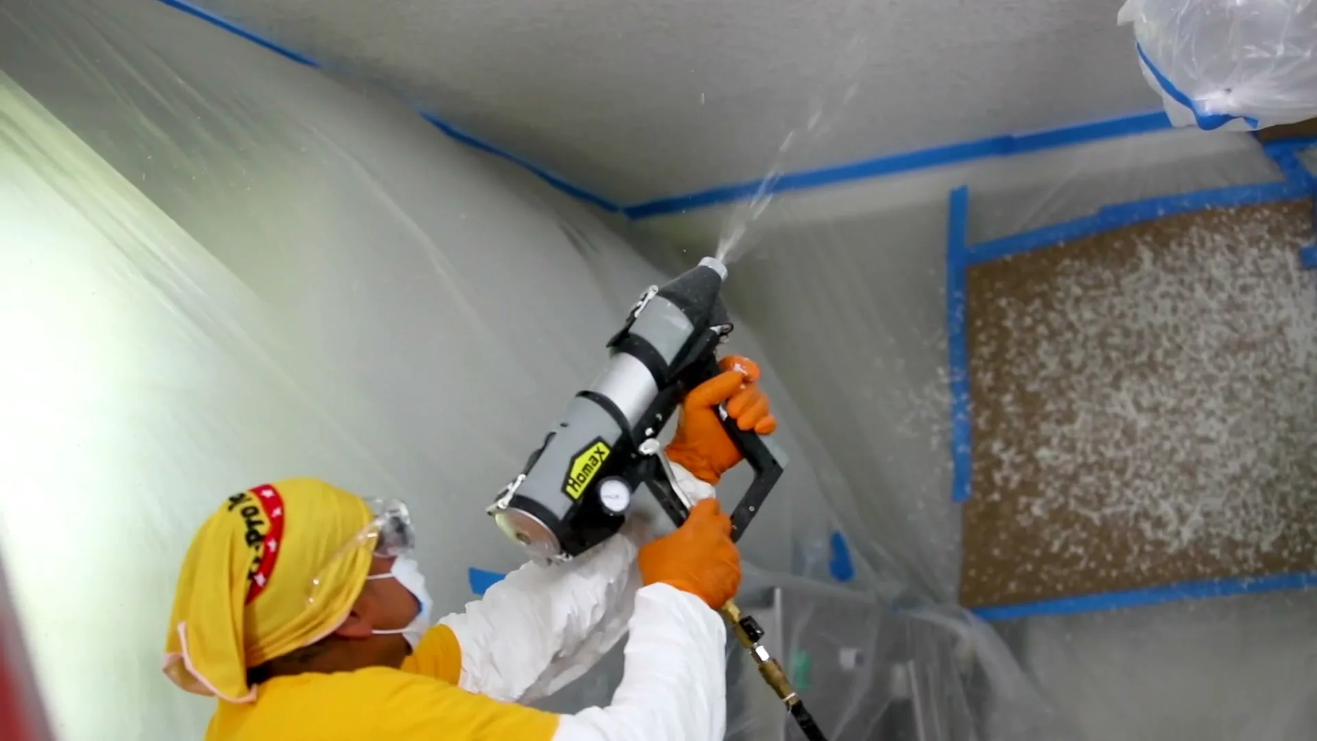 A ceiling being textured with the Homax Tex-Pro sprayer, the walls and floor under plastic.